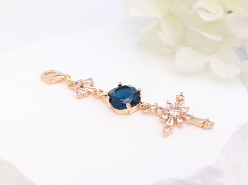 May include: A gold-toned charm featuring a lobster clasp, a small cluster of clear crystals, a central square-cut deep blue gemstone in a prong setting, and a dangling cross pendant embellished with multiple clear, baguette-cut crystals.
