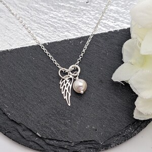 May include: A silver necklace with a delicate wing charm and a pearl pendant. The necklace is displayed on a black and silver surface, with white flowers in the background. The chain is a delicate link style.