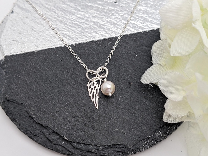 May include: A silver necklace with a delicate wing charm and a pearl pendant. The necklace is displayed on a black and silver surface, with white flowers in the background. The chain is a delicate link style.