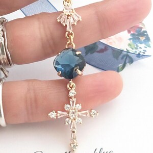 May include: A gold-toned pendant featuring a faceted dark blue cushion-cut stone, a cross-shaped charm adorned with clear baguette and round crystals, and a small fan-shaped crystal accent, all connected by delicate gold-toned metal links.