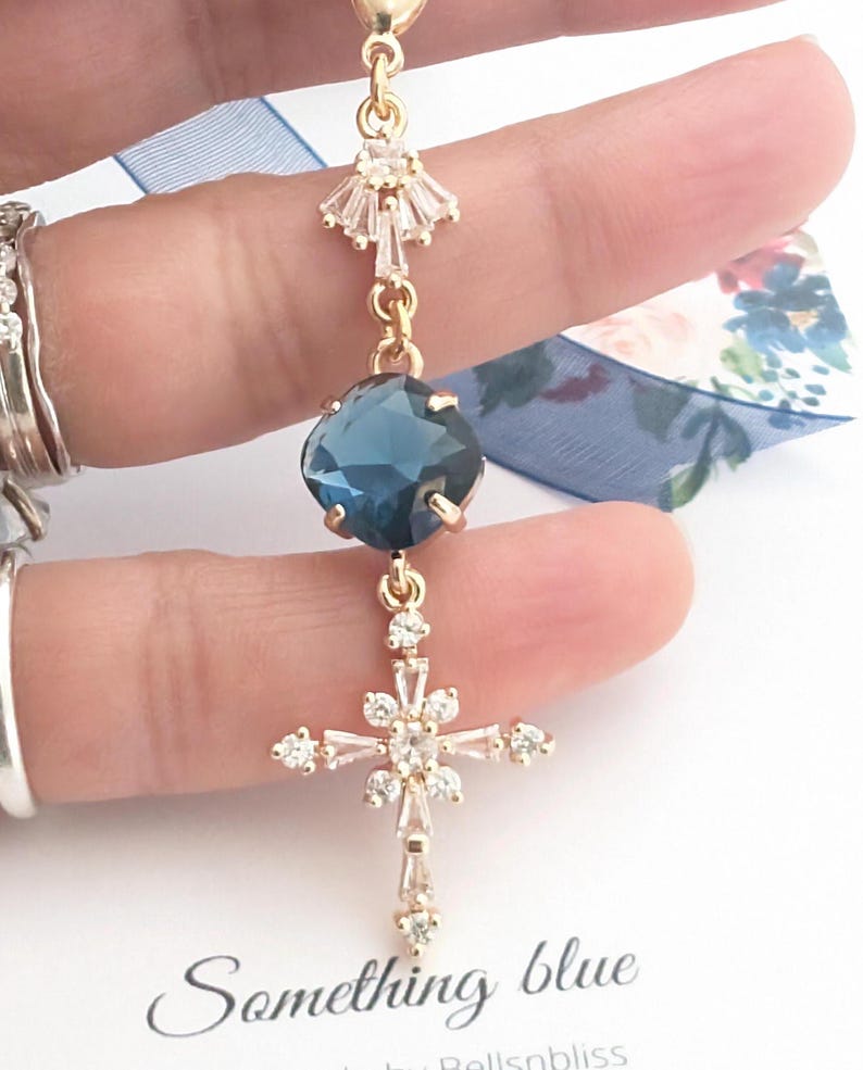 May include: A gold-toned pendant featuring a faceted dark blue cushion-cut stone, a cross-shaped charm adorned with clear baguette and round crystals, and a small fan-shaped crystal accent, all connected by delicate gold-toned metal links.