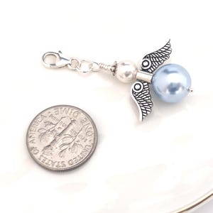 May include: A silver-toned angel charm with a light blue pearl bead body and detailed wings. The charm includes a lobster clasp and a small white pearl bead. A U.S. dime is next to the charm for size comparison.