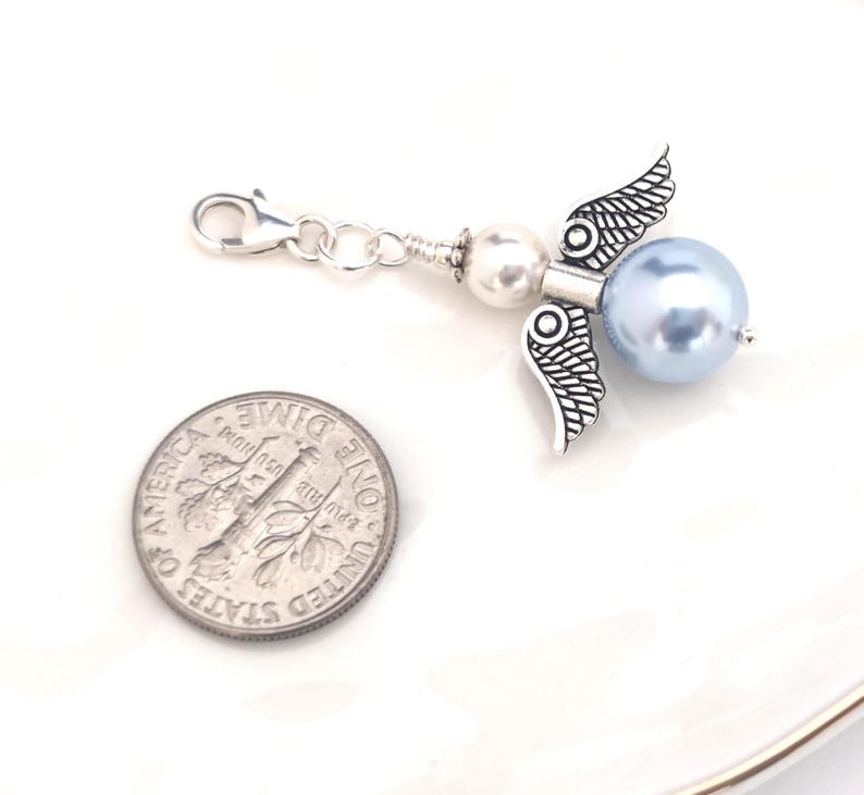 May include: A silver-toned angel charm with a light blue pearl bead body and detailed wings. The charm includes a lobster clasp and a small white pearl bead. A U.S. dime is next to the charm for size comparison.