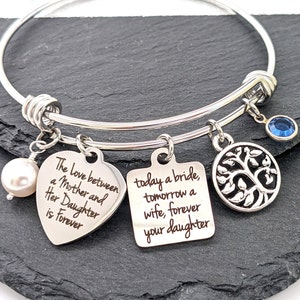 May include: Silver-toned bangle bracelet with multiple charms. Charms include a heart with the inscription "The Love between a Mother and Her Daughter is Forever", a square with text, a tree of life, a blue gem, and a pearl.
