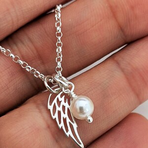 May include: A delicate silver necklace featuring a small, openwork wing charm and a lustrous white pearl pendant. The chain is a fine link style, and the necklace is shown against a neutral background.