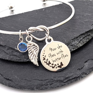 May include: Silver bangle bracelet with a knot design and a charm that says "Now she flies with butterflies". The charm has a blue crystal and a silver angel wing.