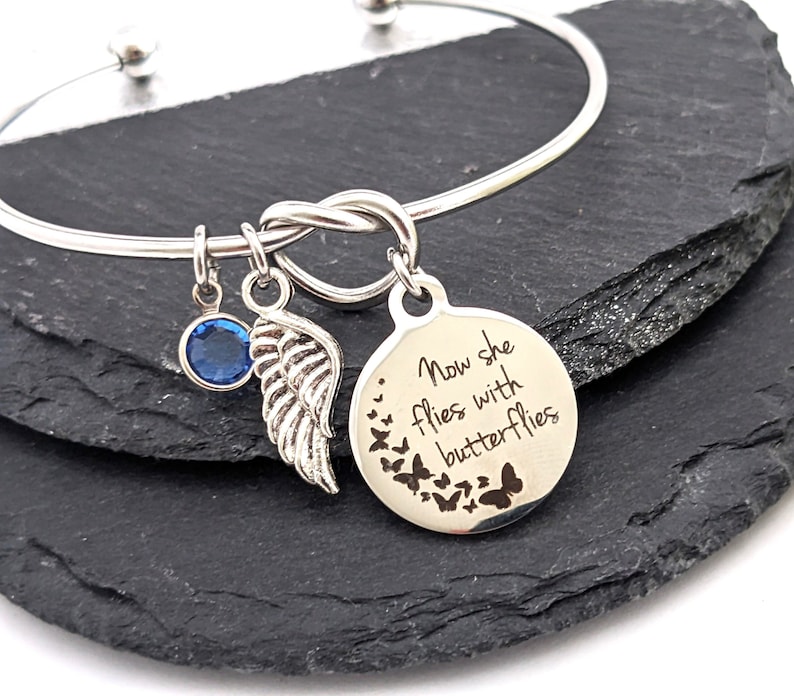 May include: Silver bangle bracelet with a knot design and a charm that says "Now she flies with butterflies". The charm has a blue crystal and a silver angel wing.