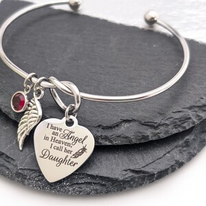 Op de afbeelding: Een zilverkleurige armband met een hartvormige bedel met de inscriptie "I have an Angel in Heaven, I call her Daughter". Het heeft ook een rode edelsteen en een veervormige bedel. Gepresenteerd op een donkere ondergrond.