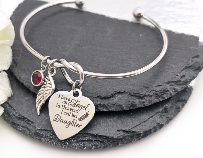 Op de afbeelding: Een zilverkleurige armband met een hartvormige bedel met de inscriptie "I have an Angel in Heaven, I call her Daughter". Het heeft ook een rode edelsteen en een veervormige bedel. Gepresenteerd op een donkere ondergrond.