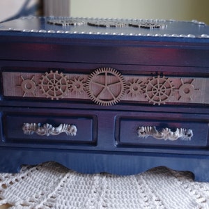 May include: A blue wooden jewelry box with three drawers and a decorative metal gear design on the front.