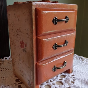 May include: A small orange wooden box with three drawers. Each drawer has a brass handle. The box has a floral design on the side.