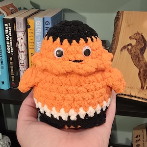 Puede incluir: Una muñeca de ganchillo naranja y negra hecha a mano con ojos saltones. La muñeca tiene una parte superior negra, una parte inferior blanca y negra y una cara sonriente. La muñeca está siendo sostenida en una mano.