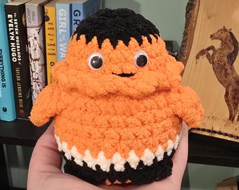 May include: A handmade orange and black crochet doll with googly eyes. The doll has a black top, a white and black bottom, and a smiling face. The doll is being held in a hand.