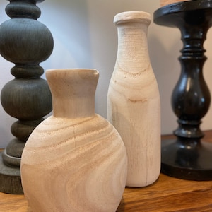 May include: Two unfinished wooden vases, one tall and slender, the other round and squat, sit on a wooden surface.