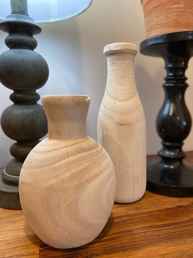 May include: Two unfinished wooden vases, one tall and slender, the other round and squat, sit on a wooden surface.