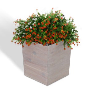 May include: A square wooden planter filled with vibrant greenery and colorful flowers. The arrangement features orange and white blossoms, set against a backdrop of green foliage and a touch of tall grass. The planter has a light gray finish.