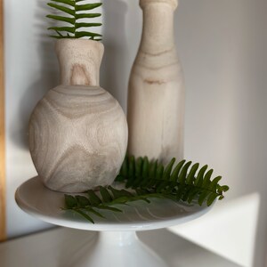 May include: Two wooden vases, one round and one tall, on a white cake stand with green fern leaves.