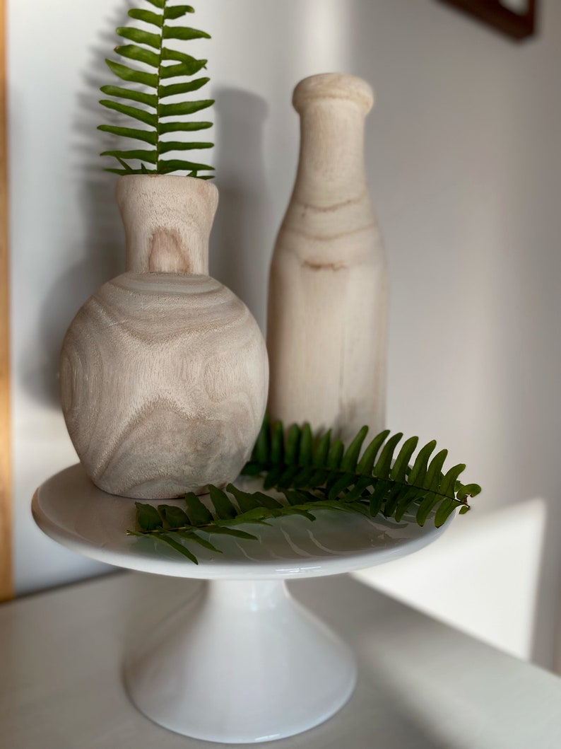 May include: Two wooden vases, one round and one tall, on a white cake stand with green fern leaves.