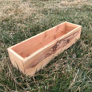 May include: A wooden planter box with a natural wood finish. The planter is made of rough-cut wood with knots and grain patterns. It is sitting on a bed of green grass.