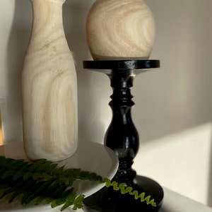 May include: Two wooden vases, one tall and slender, the other round and squat, on a black pedestal stand. The vases are both a light brown colour with a wood grain pattern. A green fern is in the foreground.