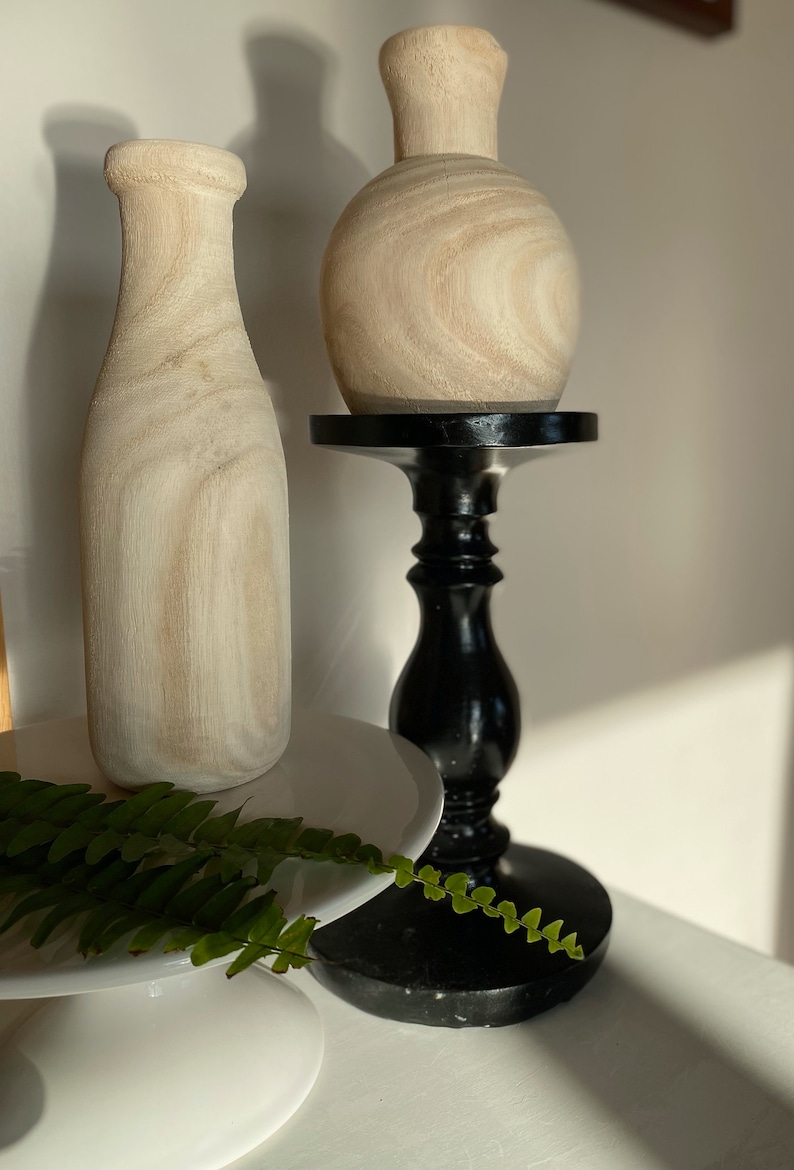 May include: Two wooden vases, one tall and slender, the other round and squat, on a black pedestal stand. The vases are both a light brown colour with a wood grain pattern. A green fern is in the foreground.