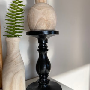 May include: A wooden vase with a round body sits on a black wooden pedestal. The vase is light brown and has a natural wood grain. The pedestal has a round base and a tall, slender column.