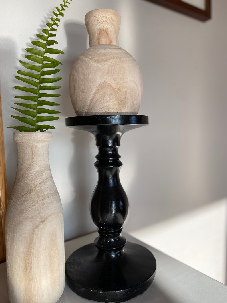 May include: A wooden vase with a round body sits on a black wooden pedestal. The vase is light brown and has a natural wood grain. The pedestal has a round base and a tall, slender column.