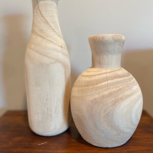 May include: Two wooden vases, one tall and narrow, the other shorter and rounder, both with a light wood grain pattern. The vases are sitting on a dark brown wooden surface.