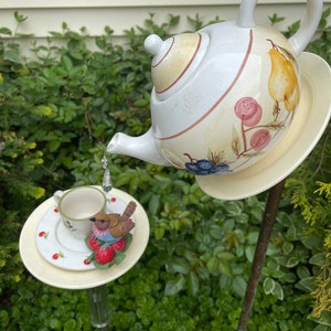 May include: A white teapot with a yellow lid and floral design pours water into a white teacup with a red bird figurine on a white saucer with red strawberries. The teapot and teacup are on metal stakes in a garden setting.