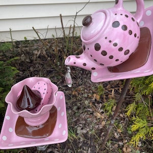 May include: Pink ceramic teapot with brown polka dots and a lid, on a pink and white polka dot plate, with a crystal hanging from the spout.  A smaller pink and white polka dot plate with a brown chocolate candy on top sits on a separate metal stake.