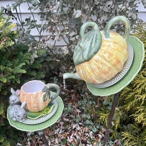 May include: A ceramic pumpkin teapot and teacup set with bunny figurines. The teapot and teacup are painted with a yellow and orange pumpkin design. The teapot has a green leaf on top. The teacup has a green leaf handle. The bunny figurines are grey and white.