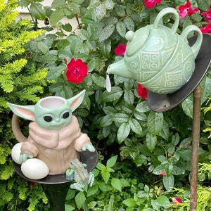 May include: A green ceramic teapot with a patterned design pours water into a brown ceramic mug shaped like a character from the Star Wars franchise. The teapot and mug are on a black stand with two white eggs and a green frog figurine.