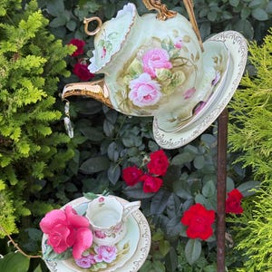 May include: A decorative garden ornament featuring a pale green porcelain teapot with pink rose patterns and a gold handle, tilted on a saucer atop a metal stake, with a matching teacup and saucer below adorned with a large pink rose sculpture.