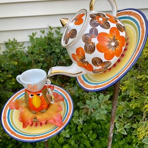 May include: A whimsical garden decoration featuring a white teapot with orange and brown floral designs, a small white teacup with a bird figurine, and two colorful plates with a blue and white border.