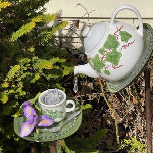 May include: A white teapot with a green ivy design pours water into a white teacup with ice cubes. The teapot and teacup are on a green glass plate with a white flower.