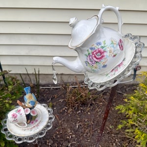 May include: A white teapot with floral designs and a gold trim is perched on a glass saucer, with a crystal hanging from the spout. A smaller teacup and saucer with floral designs are on a separate glass saucer, with a blue bird perched on the rim.