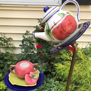 May include: A whimsical garden sculpture featuring a teapot and teacup design. The teapot is white with blue stripes and apple illustrations. A crystal prism hangs from the spout. The teacup and apple are red, set on a blue and green plate.