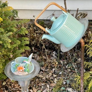 May include: A teal blue teapot with a wooden handle sits on a glass plate with a hummingbird figurine on a smaller glass plate. The teapot and plates are on a metal stand.