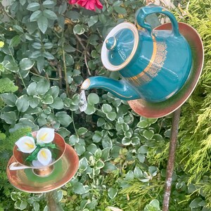 May include: A teal teapot with gold accents and a white lid is perched on a brown and green saucer. The teapot is pouring water into a white teacup with three white calla lilies inside. The teacup is on a green and gold saucer. Both are mounted on metal rods.