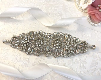 May include: A white satin ribbon with a large, ornate crystal embellishment. The embellishment is made of clear, faceted crystals arranged in a floral pattern.
