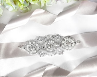 May include: A silver and white beaded sash with a floral design. The sash is on a white satin ribbon.