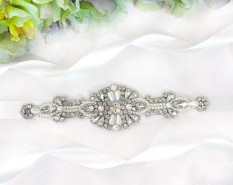 May include: A white satin ribbon with a silver and pearl embellished sash. The sash is decorated with a large, central, crystal-like stone.