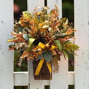 May include: A dark brown woven wicker basket filled with artificial autumn foliage, including orange and yellow flowers, green leaves, small berries, and a pinecone. A bright yellow ribbon is tied around the basket with long tails hanging down.