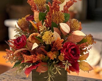 May include: A floral arrangement in a square wooden box featuring deep red roses, orange berry sprigs, yellow thistle-like flowers, and various autumn-colored leaves in shades of brown, gold, and rust, creating a dense, multi-textured display.