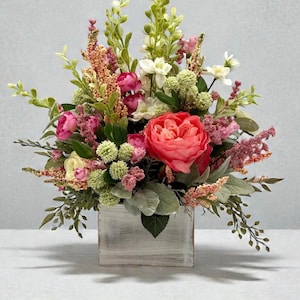 May include: A floral arrangement featuring a variety of flowers in shades of pink, red, white, and green. The flowers are arranged in a distressed, white-painted wooden box. The arrangement includes peonies, astilbe, and other greenery.