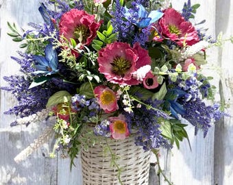 May include: A woven basket overflowing with a vibrant arrangement of artificial flowers. The bouquet features large, deep pink blooms, blue and purple accents, and green foliage. The basket hangs against a weathered white wooden fence.