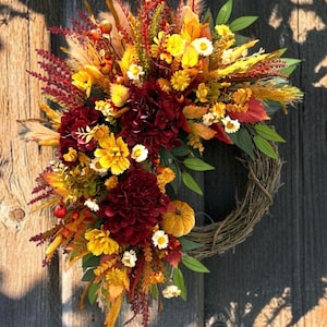 May include: A circular wreath constructed from a woven twig base, adorned with an arrangement of deep burgundy dahlias, vibrant yellow daisies, orange autumn leaves, small pumpkins, red berries, and dried grasses in shades of gold and brown.