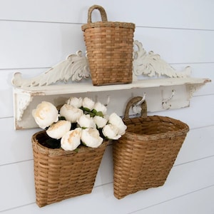 May include: A rustic, distressed white shelf with decorative carvings and three metal hooks. Two woven brown baskets hang below, one filled with white flowers. A smaller basket sits on the shelf. The baskets have rope handles.
