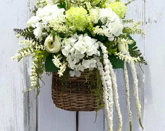 Può includere: Un cesto intrecciato traboccante di fiori bianchi e verde chiaro e verde. La composizione comprende ortensie, piccoli fiori bianchi e elementi bianchi pendenti. Il cesto è appeso su uno sfondo di legno bianco.