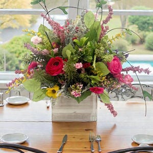 May include: A vibrant floral arrangement in a rustic wooden box. The bouquet features red, pink, yellow, and green flowers and foliage. The arrangement sits on a wooden table set with plates, silverware, and chairs.